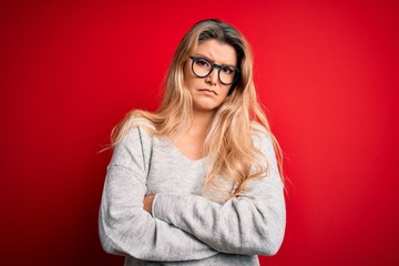 Young beautiful blonde woman wearing sweater and glasses over isolated red background skeptic and...