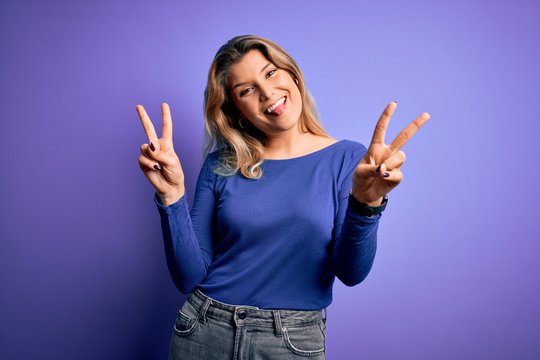 Young beautiful blonde woman wearing casual t-shirt over isolated purple background smiling with tongue out showing fingers of both hands doing victory sign. Number two.