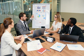 Confident bearded businessman explaining the graphs using flip chart to his multiethnic highly-skilled business partners in the office meeting room.