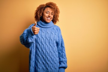 Young beautiful African American afro woman with curly hair wearing blue turtleneck sweater doing happy thumbs up gesture with hand. Approving expression looking at the camera showing success.