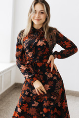 portrait of young smiling blonde slender girl in midi dress  floral pattern by a large window in light room with white wall