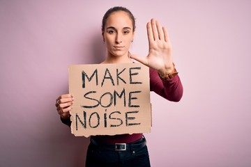 Fototapeta na wymiar Beautiful activist woman holding banner with make some noise message over pink background with open hand doing stop sign with serious and confident expression, defense gesture