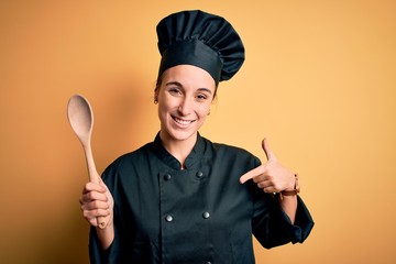Fototapeta premium Young beautiful chef woman wearing cooker uniform and hat holding wooden spoon with surprise face pointing finger to himself