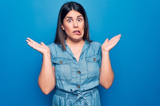 Young beautiful brunette woman wearing casual denim dress over isolated blue background clueless and confused with open arms, no idea and doubtful face.