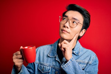 Young handsome chinese man drinking cup of coffee over isolated red background serious face thinking about question, very confused idea