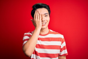 Young handsome chinese man wearing casual striped t-shirt standing over red background covering one eye with hand, confident smile on face and surprise emotion.