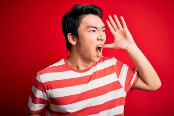 Young handsome chinese man wearing casual striped t-shirt standing over red background shouting and screaming loud to side with hand on mouth. Communication concept.