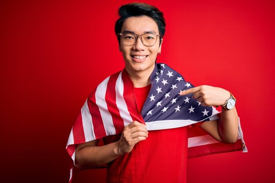 Young chinese patriotic man wearing united states of america flag over red background with surprise face pointing finger to himself