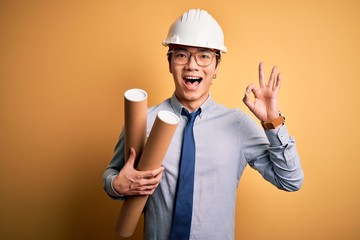 Young handsome chinese architect man wearing glasses and safety helmet holding blueprints doing ok sign with fingers, excellent symbol