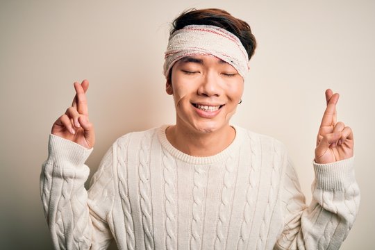 Young handsome chinese man injured for accident wearing bandage and strips on head gesturing finger crossed smiling with hope and eyes closed. Luck and superstitious concept.