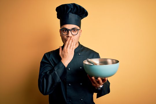 Young brazilian chef man wearing cooker uniform and hat using bowl and whisk cover mouth with hand shocked with shame for mistake, expression of fear, scared in silence, secret concept