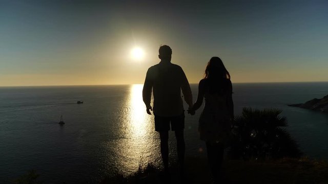 couple silhouettes stand on beach edge and kiss admiring pictorial sunset reflecting in calm ocean water in evening
