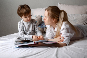 Close-up of a loving weasel mother lying with her son in bed watching a cute child reading an interesting fairy tale, a preschooler smiling at the baby boy enjoying a fairy tale before going to bed go