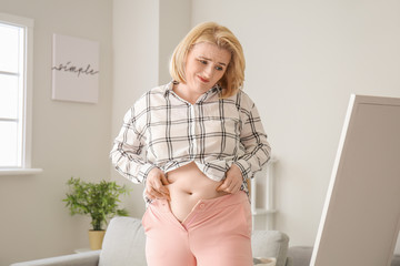 Troubled overweight woman in tight clothes near mirror at home