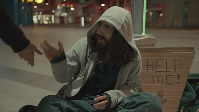 Slow motion of young bearded homeless man with long hair is threatened and beaten by a bully. 'HELP ME' sign next to him. Homeless concept. 4k.
