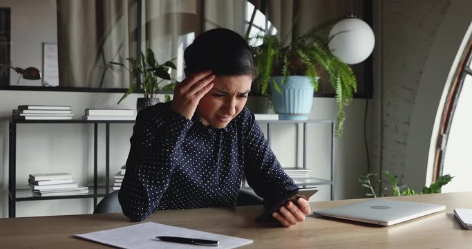 Confused unhappy indian businesswoman feels frustrated about broken phone problem in office. Annoyed female worker reading bad news in message, perplexed by mobile spam sms notification at workplace.