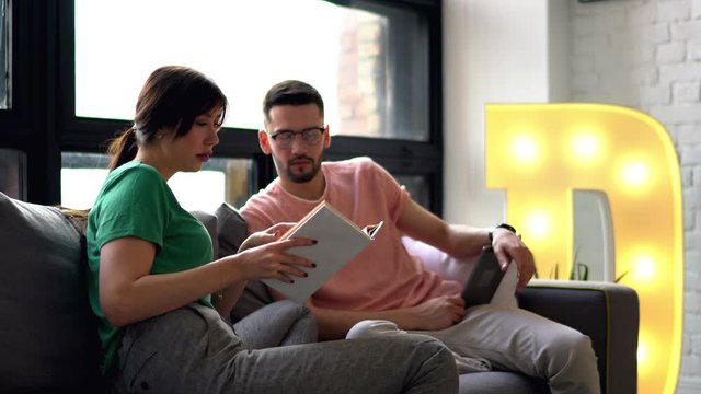 Happy couple in love discussing literature plot in bestseller and e-book relaxing on comfortable couch in modern apartment with stylish interior.Young man resting with girlfriend at home using gadget
