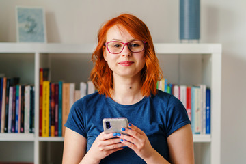 Close up portrait of young caucasian woman beautiful female natural beauty girl orange hair and blue t-shirt holding mobile smart phone at home in front of blurred bookshelf wearing eyeglasses smiling - Powered by Adobe