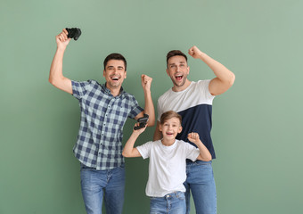 Happy gay couple with adopted child playing video game on color background