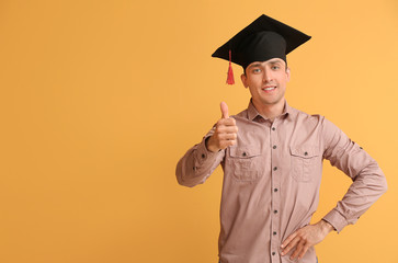 Young man in graduation hat showing thumb-up on color background