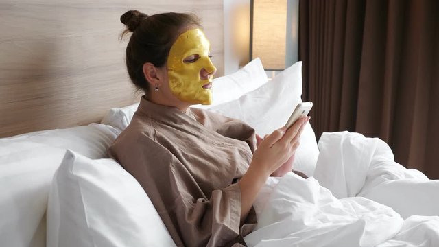 pretty woman with golden face mask uses modern smartphone to surf social nets lying in soft bed in apartment