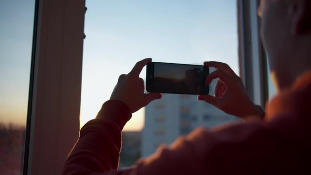 A young man is shooting a sunset on a mobile phone through a window.