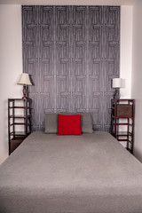 A beautiful bed in a bedroom in a modern home. An interesting wallpaper pattern serves as headboard, with gray sheets and a bright red pillow. Asian theme. Contemporary decor.