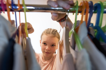 Portrait of young girl looking through wardrobe in indecision. Undecided female teenager choose clothes in cloakroom at home. A lot of dress on hangers in closet.