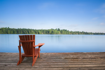 Adirondack chair sitting on a wood dock facing a calm lake. Across the water there are green trees.