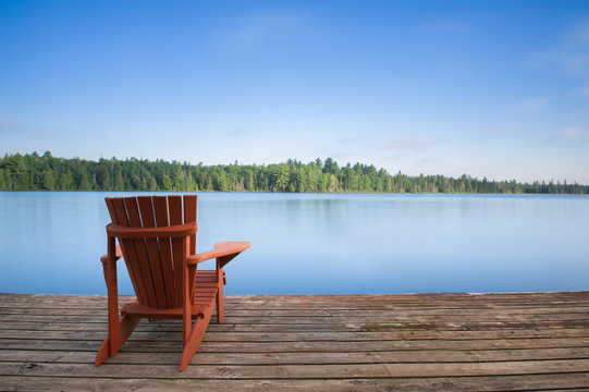 Adirondack chair sitting on a wood dock facing a calm lake. Across the water there are green trees.