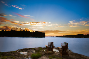 Long exposure of two Muskoka chairs sitting on a rock formation facing a calm lake at dusk in...