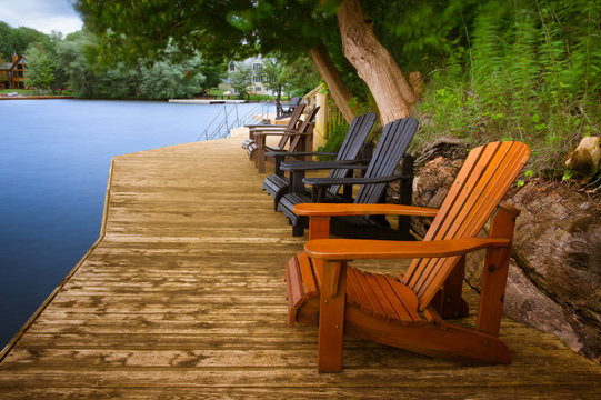 Multiple Adirondack chairs sitting on a wooden dock facing a blue calm lake. Across the water there are cottages nestled among green trees. 