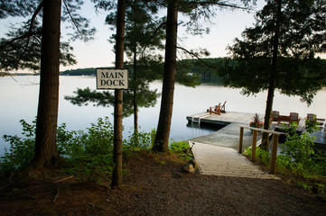 Adirondack chairs sitting on a wooden dock at sunrise. Hanging on a tree there's a sign indicating the main dock