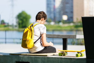 Back view of a young boy with yellow backpack and skateboard in the city park. Trendy student...