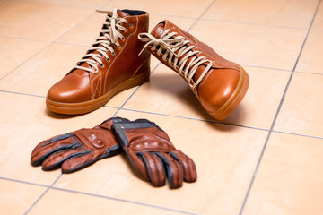 Motorcycling Relative Topics. Closeup of Protective Motorcyclist Leather Tan Sneakers Placed Indoors Along With Leather Gloves On Pale Tiles Floor.