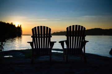 Fototapeta na wymiar Silhouette of two Adirondack chairs sit on a rock formation facing the calm waters of a lake during a sunset in Muskoka, Ontario Canada. 