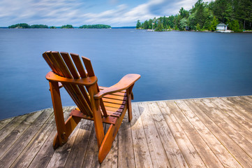 Fototapeta na wymiar Adirondack chair sitting on a wooden dock facing a blue calm lake. Across the water is a white cottage nestled between green trees. 