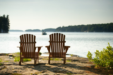 Adirondack chairs sit on a rock formation facing the waters of a lake during a sunny summer day in...