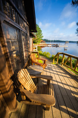 Chairs sit on a cottage wooden porch during a summer day in Canada.  A wooden dock, populated with...