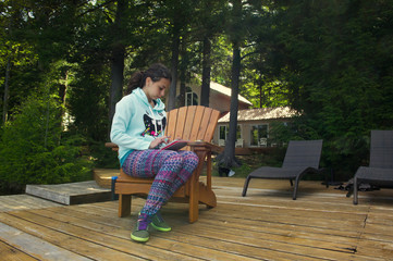 Fototapeta premium Teenager girl sit on a Muskoka chair reading on a tablet. The girl is wearing colourful clothes. A cottage nestled between trees is visible in the background.