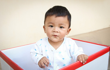 happy baby toddler sitting inside a box at home and looks at the camera. Southeast Asian boy, 7 months old.