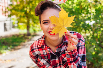 Beautiful woman with make up and hair in pin up style standing on the street, holding yellow maple leaf close to her face.