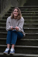 A stylish woman in a knitted sweater sits on the old city stairs and smiles at the camera holding a phone in her hands