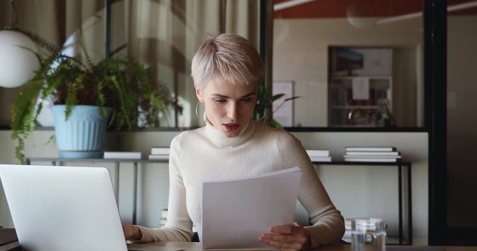 Focused young businesswoman using laptop checking paperwork doing online research. Female professional lawyer or interpreter translator working on computer analyzing documents sitting at office desk.