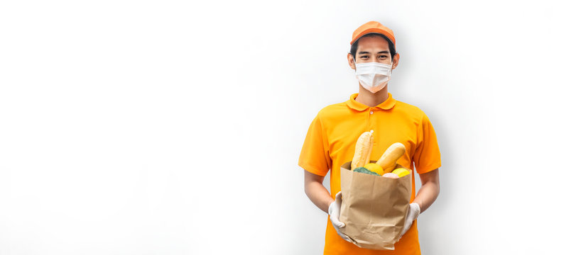 Asian Deliver Man Wearing Face Mask In Orange Uniform Holding Bag Of Food, Groceries, Fruit Standing In White Isolated Background. Postman And Express Grocery Delivery Service During Covid19 Pandemic.