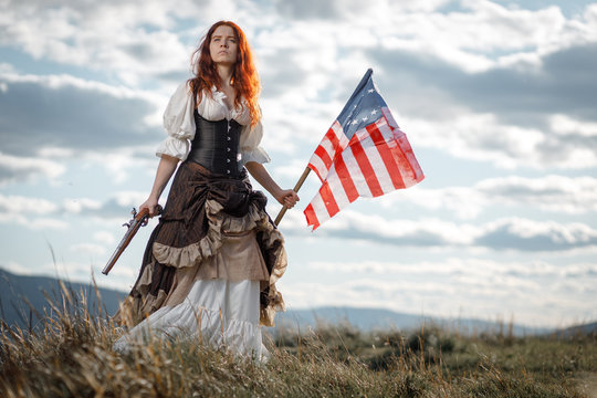 Girl in historical dress of 18th century with flag of United States. July 4 is US Independence Day. Woman of patriot freedom fighter in outdoor on background cloudy sky