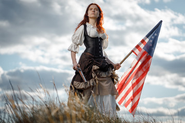Girl in historical dress of 18th century with flag of United States. July 4 is US Independence Day. Woman of patriot freedom fighter in outdoor on background cloudy sky - obrazy, fototapety, plakaty