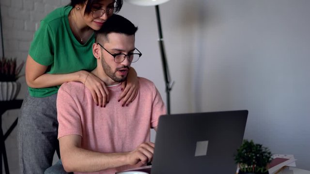 Caring young woman with cool tattoo on hand hugging lovely husband and help him to discuss design sketch during work at modern laptop computer at home interior on weekend 
