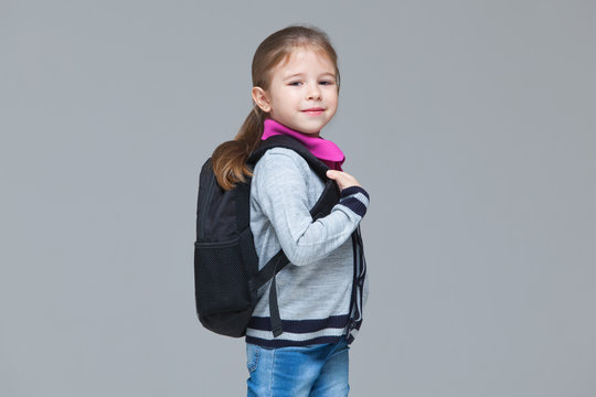 Primary School Girl In Jeans And Uniform Jacket Is Wearing Backpack. Isolated On Grey Background