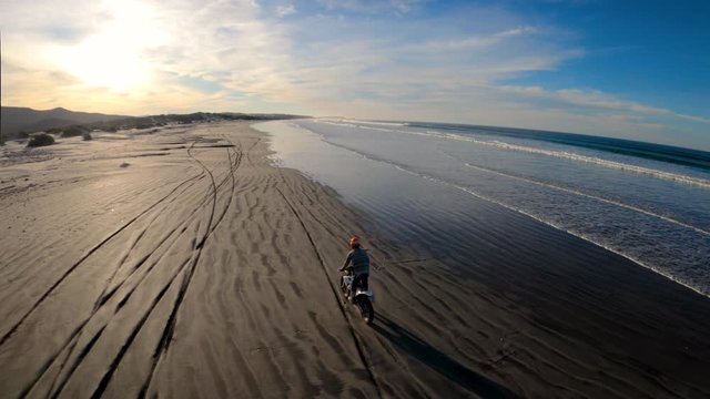 Wild FPV Race Drone Drifting Behind Motorcycle Stuntman on Beach at Sunrise
